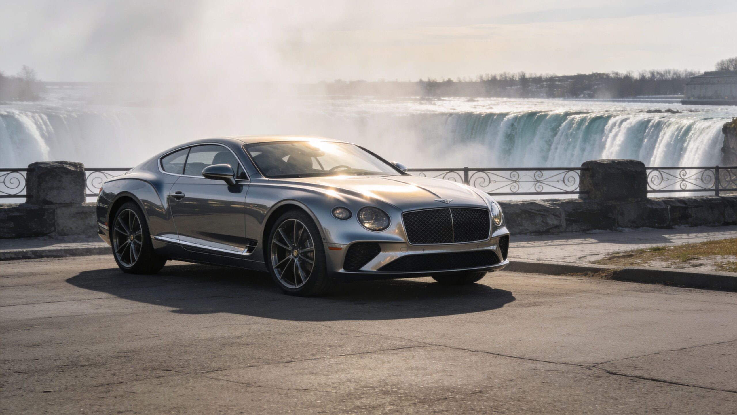 A luxurious silver Bentley coupe parked on a paved road with Niagara Falls in the background.