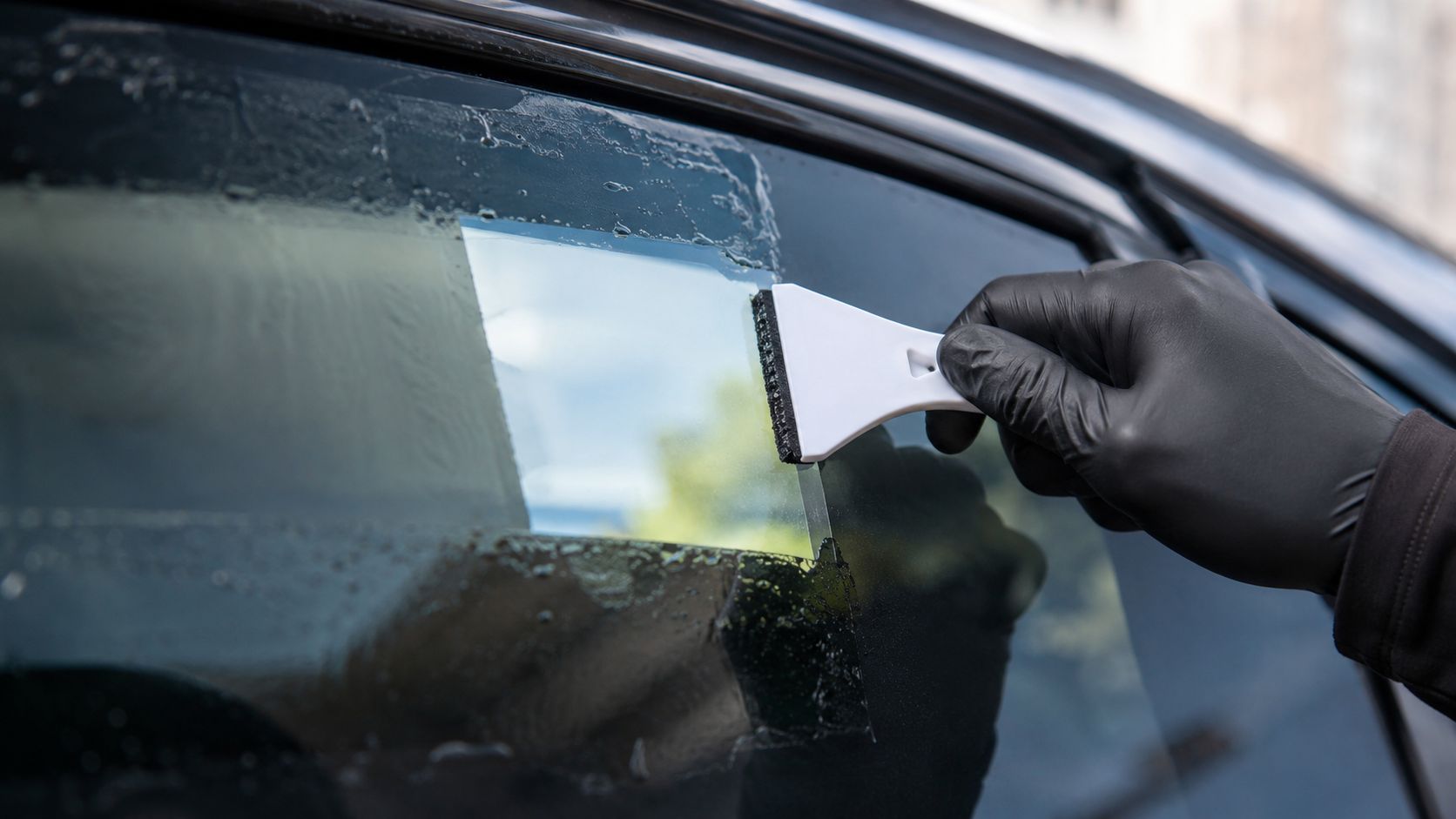 A professional technician wearing black gloves using a plastic squeegee to remove old window tint from a car.
