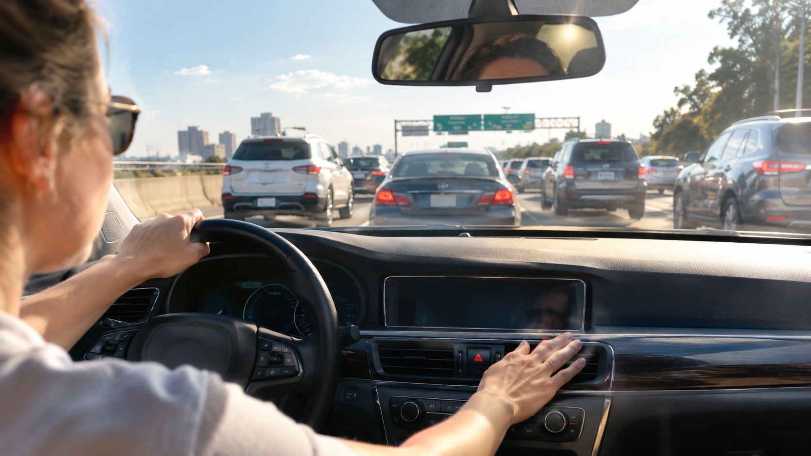 A woman drives a car on a highway while touching the dashboard controls of the vehicle.